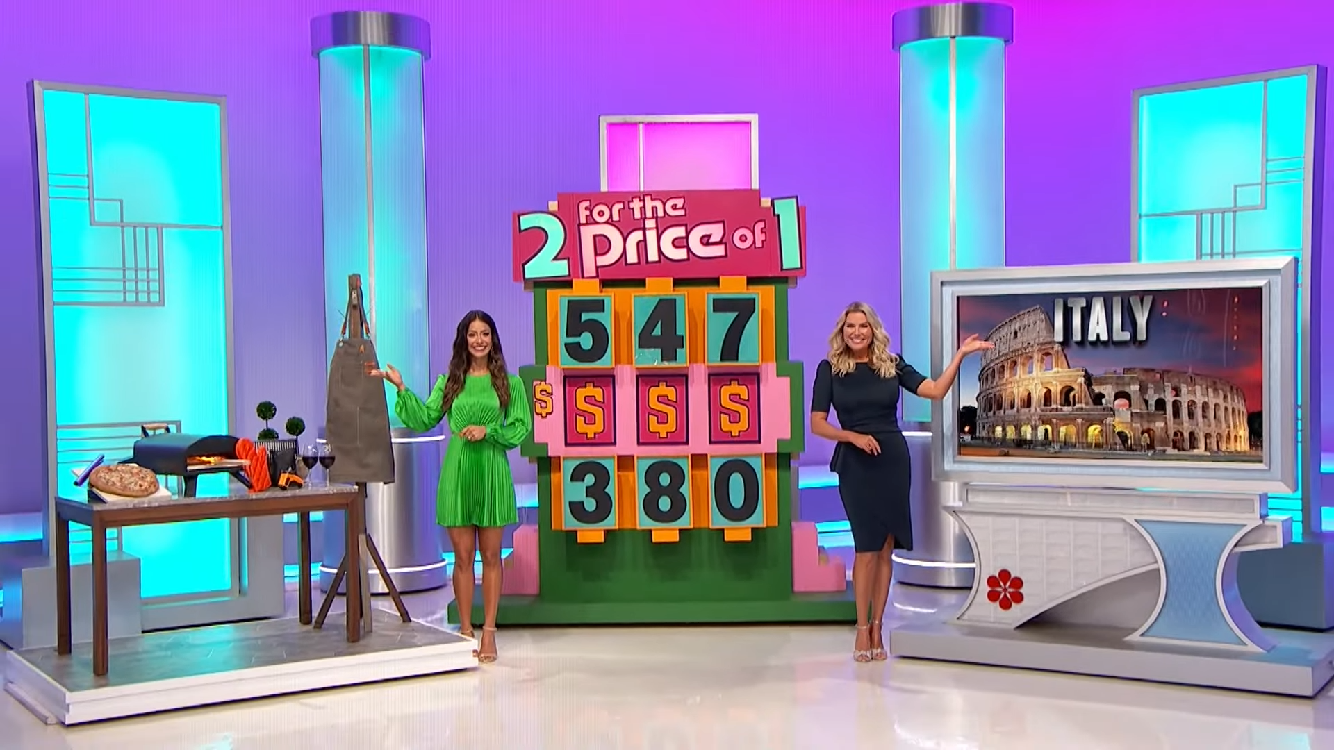 Two women stand beside a colorful game board labeled '2 for the Price of 1', with numbers 547 and 380 displayed. The background features a digital image of the Colosseum in Italy.