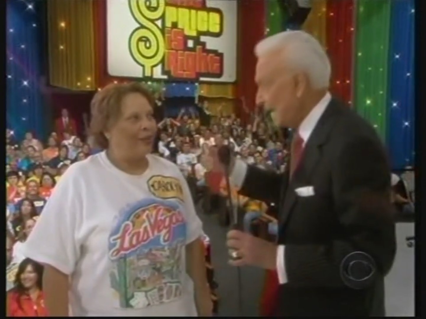 A contestant in a Las Vegas-themed shirt participates on stage with a host during a game show. A colorful backdrop and a lively audience are visible.
