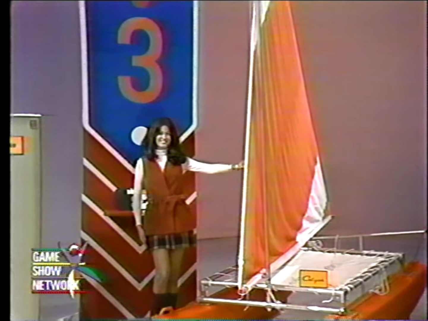 A contestant stands next to a catamaran showcasing its colorful sails during the game 'Most Expensive.' The backdrop features a large number 3.