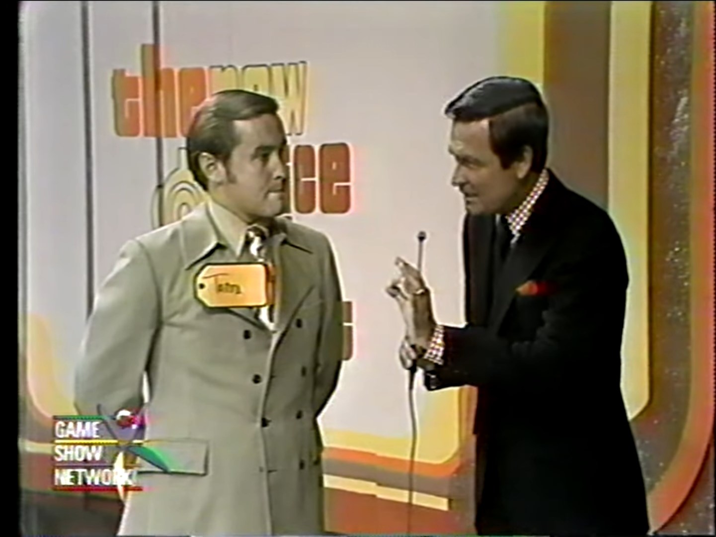 A game show host speaks to a contestant named Tom on stage of The Price Is Right, with colorful backdrop and game show elements visible.