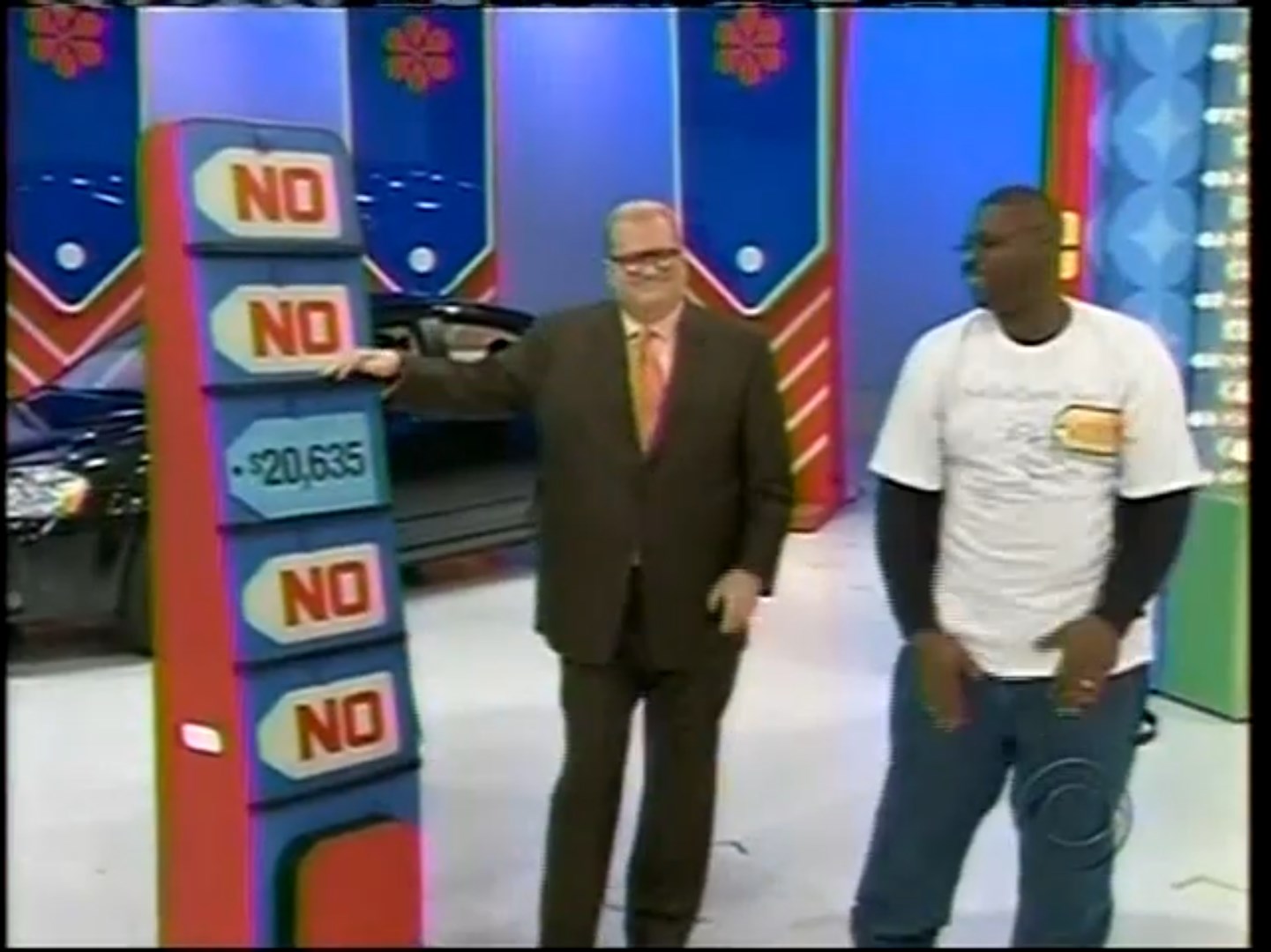 Drew Carey and host standing next to a game display with five price tags, and a car in the background.