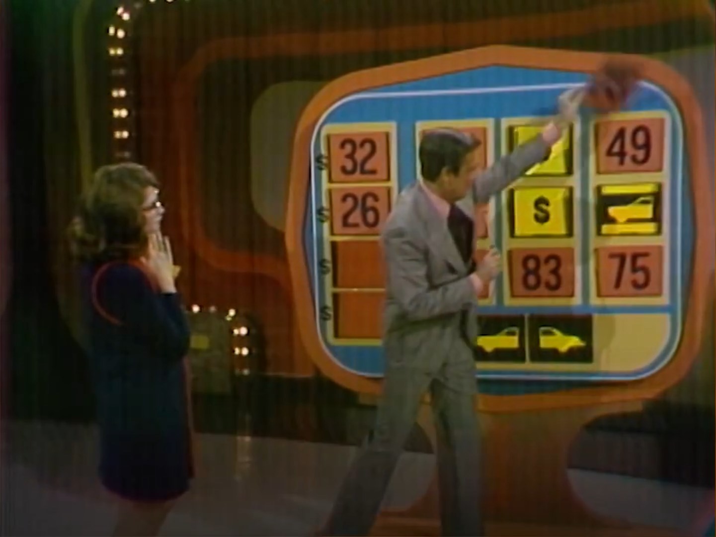 A game show contestant looks on anxiously as the host reveals numbers on a Money Game board in 1972, with several dollar signs indicating incorrect guesses. The setting features colorful lights and a retro game show aesthetic.