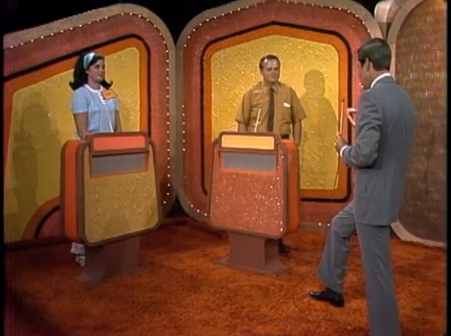 Two contestants stand behind podiums on a game show set, with a host gesturing towards them.