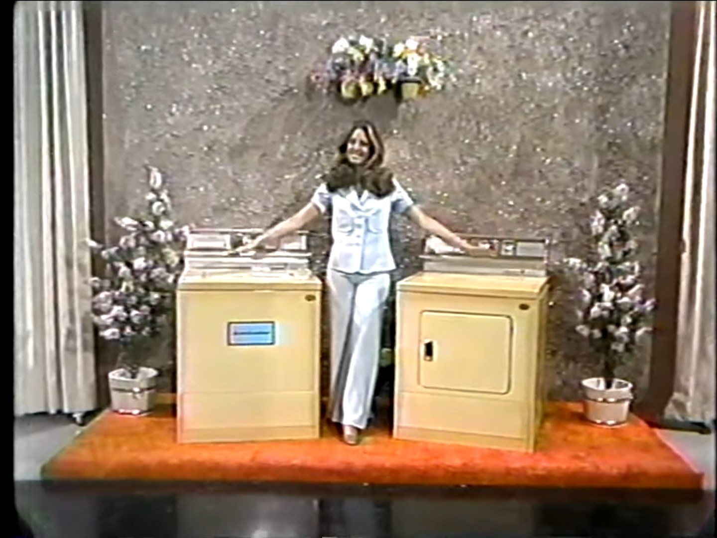 A model stands between a washing machine and dryer, smiling and gesturing towards them, with a sparkly backdrop and decorative plants in the background.