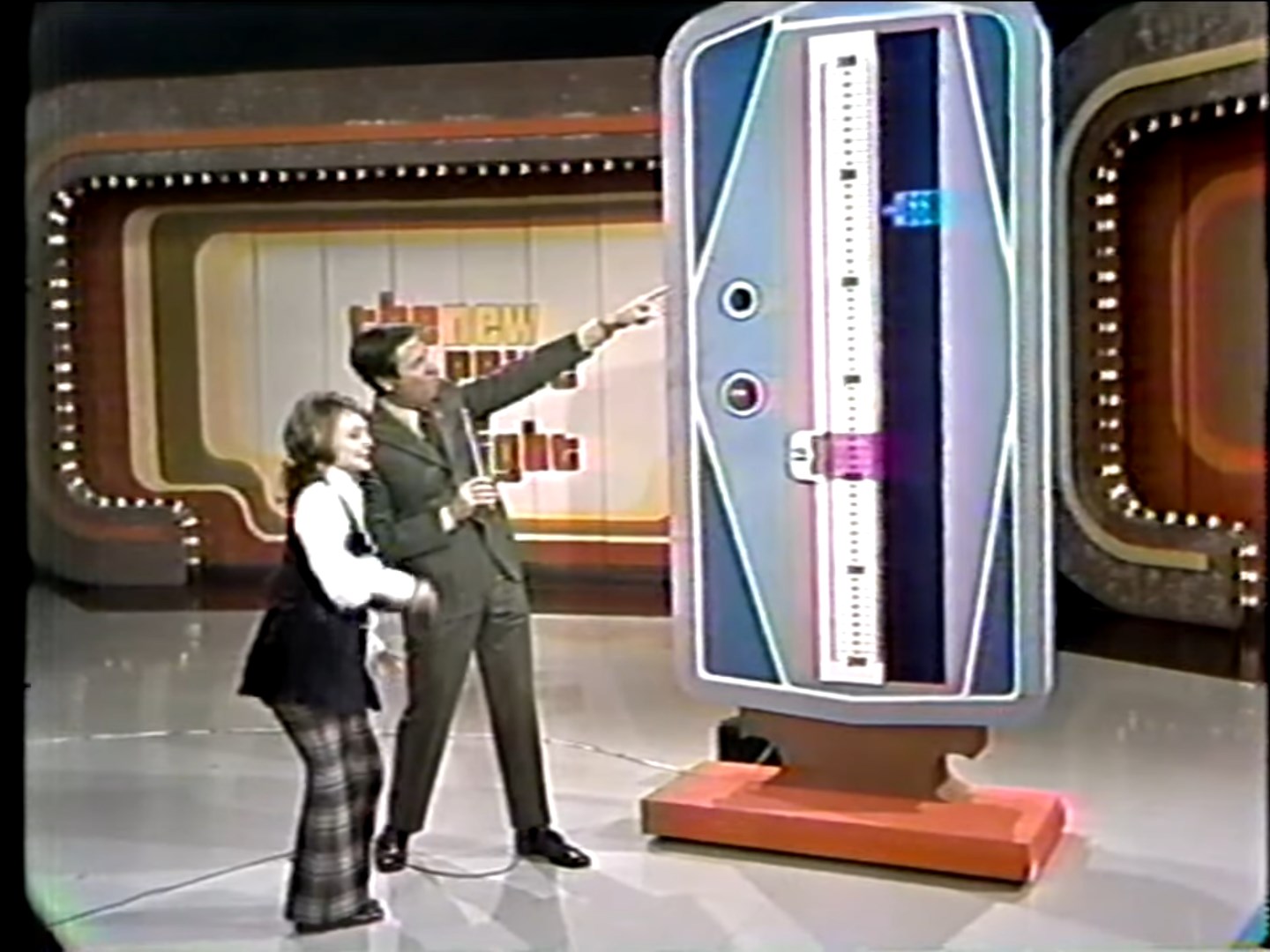 A contestant and a host stand in front of the Range Game device, with the host pointing while the contestant prepares to participate.