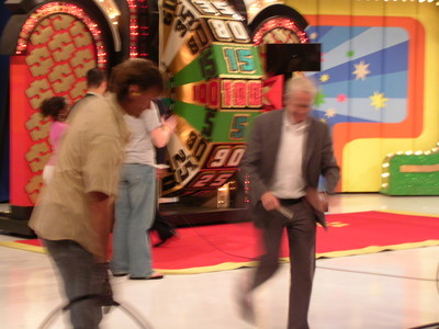 A blurred scene from backstage at a game show, featuring a man in a suit and another person partially visible, with a large spinning wheel and colorful set elements in the background.