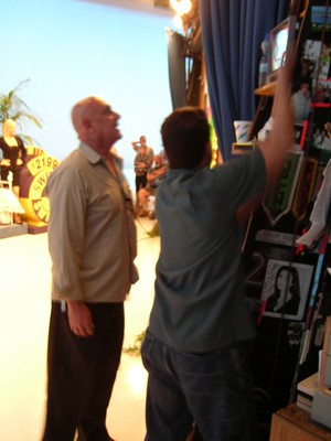 Two men backstage on a television set, one observing and the other adjusting items on a board.