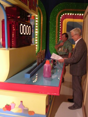 Two people backstage at a colorful game station on a television set, with one man reading a sheet of paper while the other stands near him.