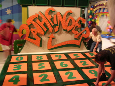 A game show set featuring a large sign that reads 'Pathfinder' with colorful orange letters, surrounded by colorful decorations. Several crew members are adjusting orange tiles on the green game board.