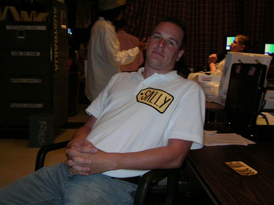 A man sitting casually in a chair, wearing a shirt with the name tag 'SALLY,' in a backstage setting with blurred figures and equipment in the background.