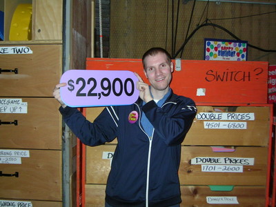 A contestant backstage at a game show holding a sign with the amount $22,900, smiling in front of storage boxes labeled with prices.