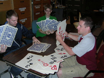 Three people sitting around a table with large playing cards, one holding a card showing the number 6, another displaying other cards, and the third person engaging with cards in their hands.