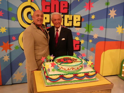 Two men posing in front of a colorful backdrop celebrating a special occasion, with a decorated cake on a table.