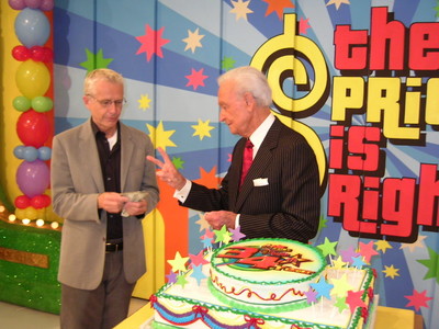 A man in a suit hands a piece of paper to another man in a black suit, standing next to a colorful cake decorated for The Price Is Right.