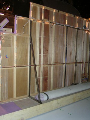 Backstage view of a wooden wall structure with lights and a rolled carpet on the floor.