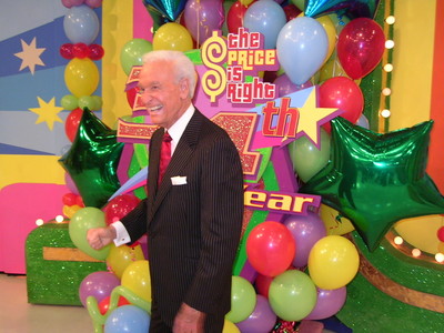 A cheerful host stands in front of a colorful backdrop with balloons and decorations celebrating the 34th anniversary of a game show.