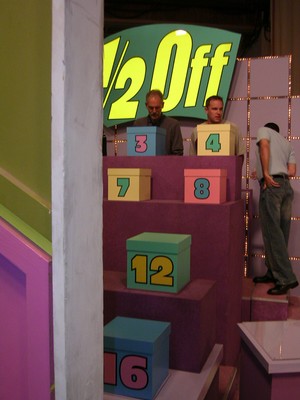 Contestants preparing for the game 'Half Off' on The Price Is Right set, with colorful blocks numbered 3, 7, 8, 12, and 16 in view.
