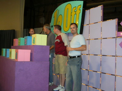 Contestants standing near colorful game boxes on a set for a game show, with a sign reading '1% Off' in the background.