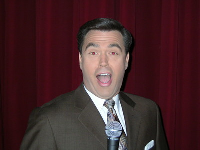 A man in a suit smiling widely and holding a microphone in front of a red curtain.