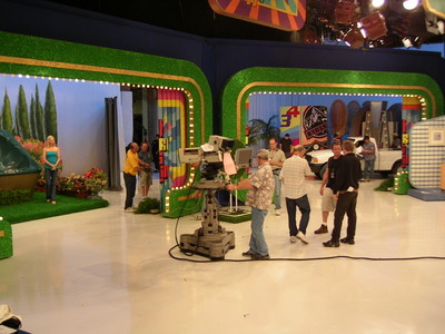 Behind the scenes at the Season 34 premiere of a TV show, featuring crew members preparing set elements, including a camera, colorful backdrops, and props.