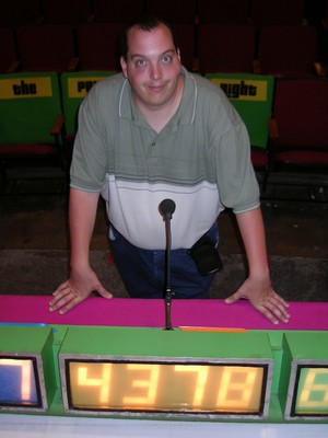 A person standing at a game show podium with a microphone, smiling at the camera, with colorful seating in the background.