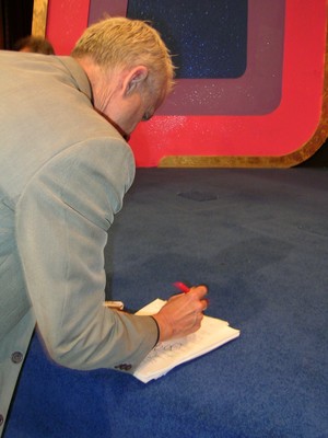 A person in a suit is writing on a notepad while standing on a blue carpet stage with a colorful background.