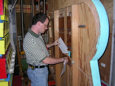 A person wearing a striped shirt is adjusting paper on a wooden structure backstage, surrounded by colorful game elements.