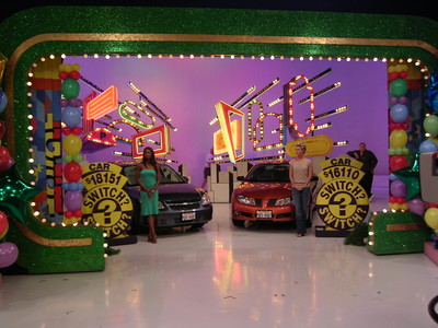 Two contestants stand in front of colorful game show set with cars and bright lights, featuring the text 'SWITCH?' on signs.