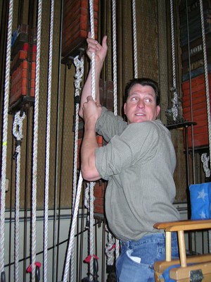 A person is pulling on a rope in a backstage setting, surrounded by rigging equipment and ropes.