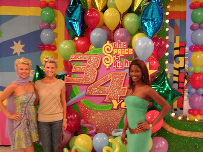Three women posing in front of a colorful backdrop celebrating the 34th anniversary of 'The Price is Right,' adorned with balloons and a large sign.