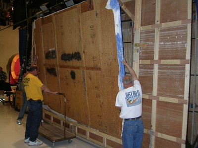 Two crew members adjusting a large wall panel backstage at a television studio.