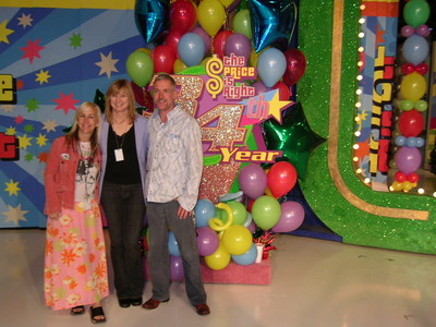 Three individuals posing together in front of a colorful backdrop adorned with balloons and decorations celebrating the 34th year of 'The Price is Right.'