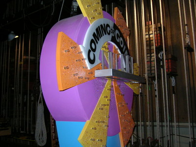 A colorful studio prop featuring a large circular structure with the text 'COMING OR GOING' prominently displayed, surrounded by vibrant yellow and orange elements. The backdrop includes studio rigging equipment.