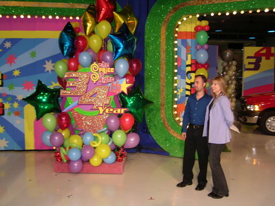 A colorful backdrop featuring balloons and stars celebrates the 34th year of 'The Price is Right', with two individuals posing for a photo in front of it.