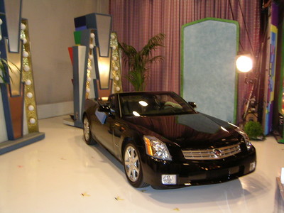 Black convertible car displayed on a brightly lit stage with colorful decorations in the background.
