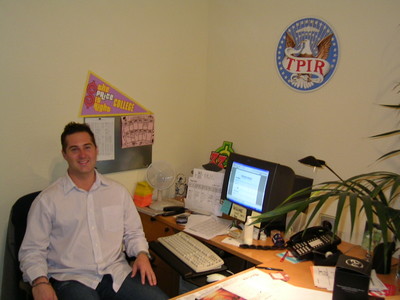 Scott Schalk sitting in an office chair at a desk with a computer, paperwork, and a decorative wall plaque reading 'TPIR'.