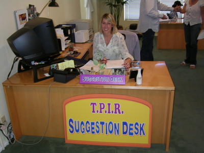 Vanessa Voss sitting at a desk labeled 'T.P.I.R. SUGGESTION DESK' with a computer and paperwork in a brightly lit room.