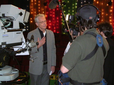 A man in a gray blazer speaks to a camera crew backstage, surrounded by colorful lights and studio equipment.