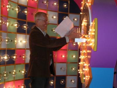 A man in a brown jacket holding a script stands next to a brightly colored game show wheel, surrounded by lights and vibrant background panels.