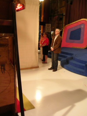 Two individuals stand backstage on a television set, with one wearing a red shirt and the other in a brown jacket. The set features colorful geometric shapes and blue stairs.