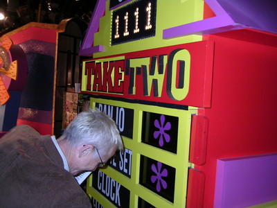A person examining a brightly colored game show board labeled 'TAKE TWO' with additional text and graphics, set in a backstage environment.