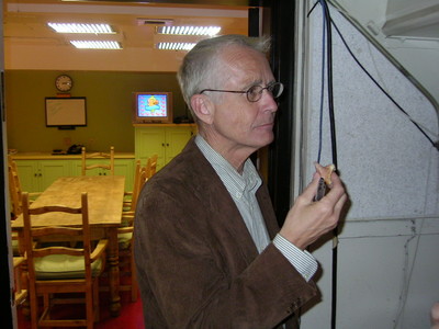 A man in a brown jacket is holding a snack while standing in a brightly lit room with a dining table and chairs in the background.