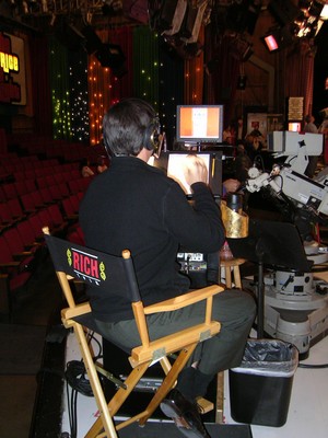 A behind-the-scenes view of a television set during a taping. The image shows Rich Fields seated in a director's chair with 'RICH' on the back, using equipment and looking at a screen. In the background, there are empty audience seats and colorful stage decorations.