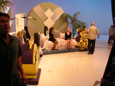 Behind the scenes of a game show set featuring two women seated on a couch, surrounded by game elements, with a production team member in the foreground.