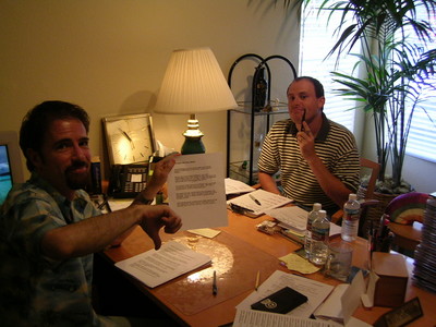 Stan Blits and Adam Sandler sitting at a table covered with papers and office supplies, one giving a thumbs down gesture while holding a document.