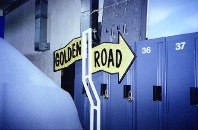 A directional sign pointing to 'Golden Road' in front of lockers, illustrating props from TV gameshow.
