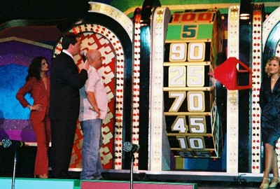 A game show host interacts with a contestant next to a large spinning wheel marked with numbers, while a female contestant stands in the background.