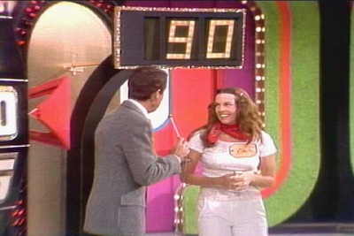 A game show host interacts with a smiling contestant, showing a score of 90 on a large display in the background.