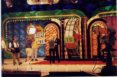 A scene from 'The Price Is Right' game show featuring a large wheel, a host presenting, and camera crew members in the background.
