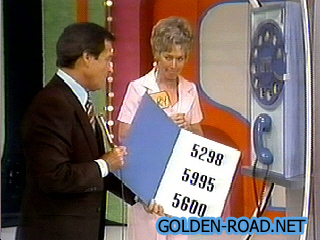 A game show host and a female contestant stand next to a large payphone, holding a sign displaying three phone numbers: 5298, 5995, and 5600.